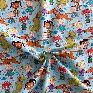 May include: A blue fabric with a repeating pattern of cartoon characters from the Nickelodeon show Rugrats. The characters include Tommy Pickles, Chuckie Finster, Phil and Lil DeVille, Angelica Pickles, and Susie Carmichael. The characters are surrounded by stars and planets.