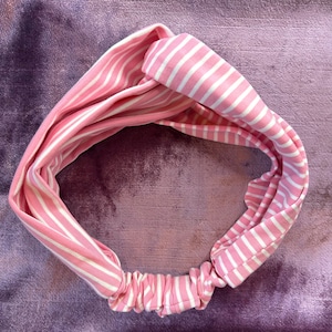 May include: A pink and white striped headband with a knotted design. The headband is made of a soft, stretchy fabric.