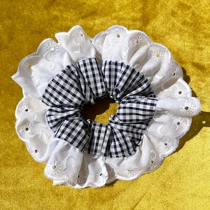 May include: A white hair scrunchie with a black and white gingham pattern. The scrunchie has a scalloped edge with white embroidery.