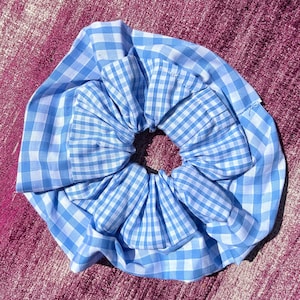 Puede incluir: Un coletero de pelo de vichy azul y blanco. El coletero está hecho de dos capas de tela y se reúne en el centro para crear una forma de flor.