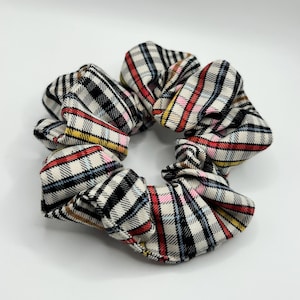 May include: A plaid scrunchie in cream, black, red, pink, yellow, and blue. The fabric is patterned with a classic check design. This hair accessory is a stylish addition to any hairstyle.