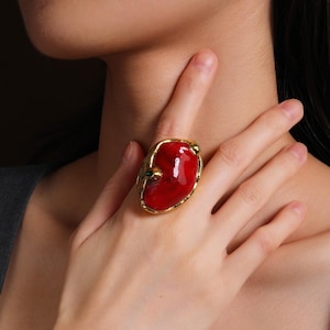 May include: A gold-toned ring featuring a large, oval-shaped red stone. The ring has a textured gold band and small green accents. The ring is worn on a finger, with the hand and part of the person's neck visible.