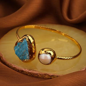 May include: Gold-coloured open cuff bracelet with a turquoise stone and a pearl. The bracelet has a hammered texture and rests on a yellow-toned surface, against a brown fabric backdrop.