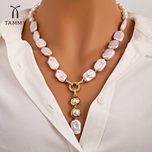 May include: A white baroque pearl necklace with a gold pendant featuring two pearls and a circle of sparkling stones. The necklace is on a model wearing a white shirt.