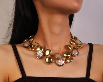May include: A gold-toned statement necklace featuring large, irregular, circular pendants. Each pendant has a textured gold border and a pearlescent, off-white center. The necklace is worn around the neck, showcasing its layered design.