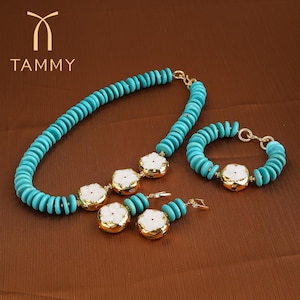 May include: A jewelry set featuring a turquoise beaded necklace, bracelet, and earrings. The set includes gold-toned accents and white flower-shaped pendants. The jewelry is displayed on a brown surface.