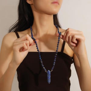 May include: A long necklace featuring dark blue lapis lazuli beads and a faceted lapis lazuli pendant. The necklace is held up, showcasing its length and design. The beads are small and round, with a silver-colored metal accent.