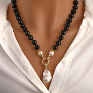Könnte beinhalten: Eine Halskette aus schwarzen Onyxperlen mit einem goldenen Anhänger, der eine große Barockperle und kleinere Perlen trägt. Der Anhänger hat ein kreisförmiges Design mit kleinen Diamanten.