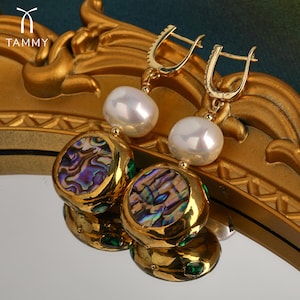 May include: Gold earrings with a pearl and abalone shell design. The earrings have a gold hoop and a pearl dangling from it. Below the pearl is a round abalone shell with green gemstones around the edge.