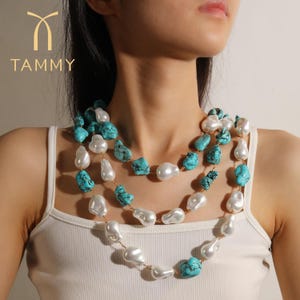 Può includere: Collana a tre fili con grandi perle bianche irregolari e pietre turchesi. La collana è infilata su una catena color oro. Il marchio "TAMMY" è visibile in alto a sinistra.