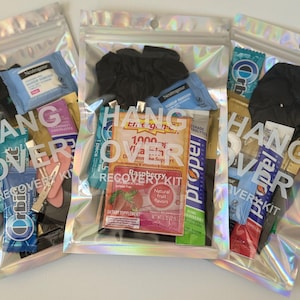 May include: Three iridescent zipper bags, each labelled "Hangover Recovery Kit". The bags contain items like makeup remover wipes, hair ties, and supplement sachets. The bags are clear, allowing the contents to be seen.
