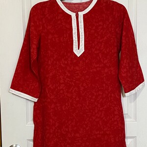 Könnte beinhalten: Eine rote Tunika mit weißem Besatz an Ausschnitt und Ärmeln. Die Tunika hat einen kleinen V-Ausschnitt und eine lockere Passform.