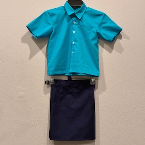 May include: A turquoise short-sleeved button-down shirt with a chest pocket and a navy blue skirt, hanging on a hanger. The shirt and skirt are made of a smooth fabric, likely cotton or a cotton blend. The outfit is suitable for school or a uniform.