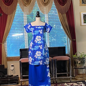May include: A blue dress with a floral pattern, displayed on a mannequin. The dress features a square neckline and short sleeves. The floral design includes white hibiscus flowers and blue leaves. The skirt is a solid blue color. The dress is a traditional style.