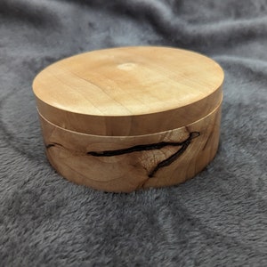 May include: A small, round wooden box with a lid. The box is made of light wood with dark brown streaks. The lid is smooth and has a natural finish.