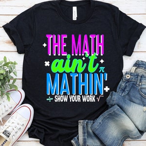 May include: Black t-shirt with the text "THE MATH ain't MATHIN'" in pink, green, and blue. The text "SHOW YOUR WORK" is below in white with a division symbol and a square root symbol.