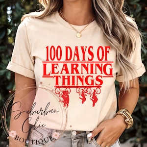 May include: A cream-coloured t-shirt featuring the red text "100 Days of Learning Things" and red bicycle graphics. The shirt has a round neck and short sleeves. A gold necklace and bracelet are also visible in the image.