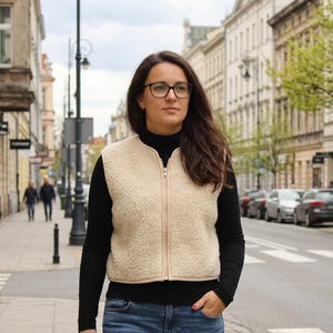 Op de afbeelding: Een crèmekleurig, mouwloos vest met ritssluiting, gemaakt van een gestructureerd, fleece-achtig materiaal. Het vest wordt gedragen over een zwart shirt met lange mouwen en een blauwe spijkerbroek. Het kledingstuk heeft een eenvoudige, kraagloze hals.