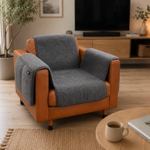 May include: A tan leather armchair featuring a dark gray, textured fabric cover that drapes over the back, seat, and armrests. The cover has a small rectangular tag on the left armrest and is designed to protect the surface of the chair.