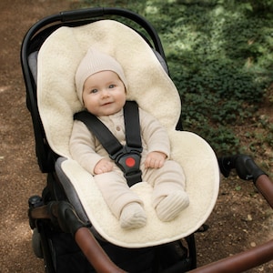 May include: A cream-colored, plush sheepskin stroller liner featuring a contoured shape with cutouts for a five-point harness system. The liner is positioned inside a black stroller, providing a soft, textured surface for a seated infant.