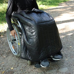 Könnte beinhalten: Eine schwarze, gesteppte Rollstuhl-Abdeckung. Die Abdeckung dient dem Schutz und der Wärme. Sie besteht aus einem glänzenden, wasserabweisenden Material. Die Abdeckung ist so konzipiert, dass sie über den Schoß und die Beine einer Person im Rollstuhl passt.