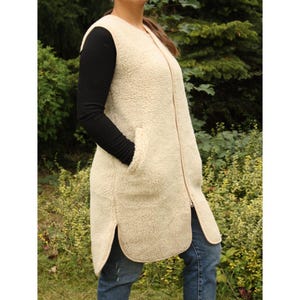 Può includere: Un gilet lungo senza maniche realizzato in materiale pile sherpa testurizzato color crema. Il capo presenta una chiusura frontale con cerniera a tutta lunghezza, uno scollo rotondo e due tasche laterali con orli arrotondati sul fondo.
