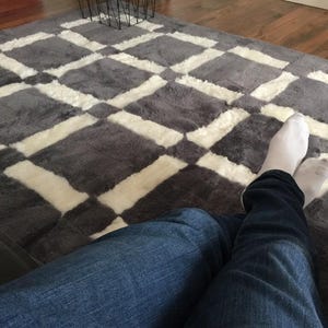 May include: A person wearing blue jeans and white socks sits on a grey and white chequered rug. The rug has a soft, fluffy texture.
