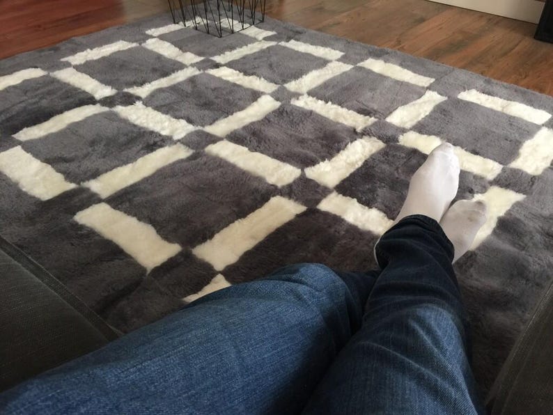 May include: A person wearing blue jeans and white socks sits on a grey and white chequered rug. The rug has a soft, fluffy texture.