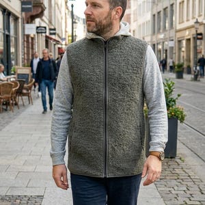 Könnte beinhalten: Eine Reißverschlussweste aus olivgrünem, strukturiertem Fleece, getragen über einem hellgrauen Kapuzenpullover mit langen Ärmeln. Die Weste verfügt über einen durchgehenden Frontreißverschluss, einen Stehkragen und ein ärmelloses Design.