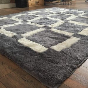 May include: A grey and white sheepskin rug with a geometric pattern. The rug has a soft, fluffy texture.