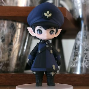Könnte beinhalten: Eine kleine Spielfigur mit spitzen Ohren, die eine dunkelblaue Militärmütze mit einem hellen Blattsymbol, einen passenden dunkelblau-schwarzen Mantel mit kleinen sternförmigen Knöpfen und dunkle Stiefel trägt und auf einem runden Sockel steht.