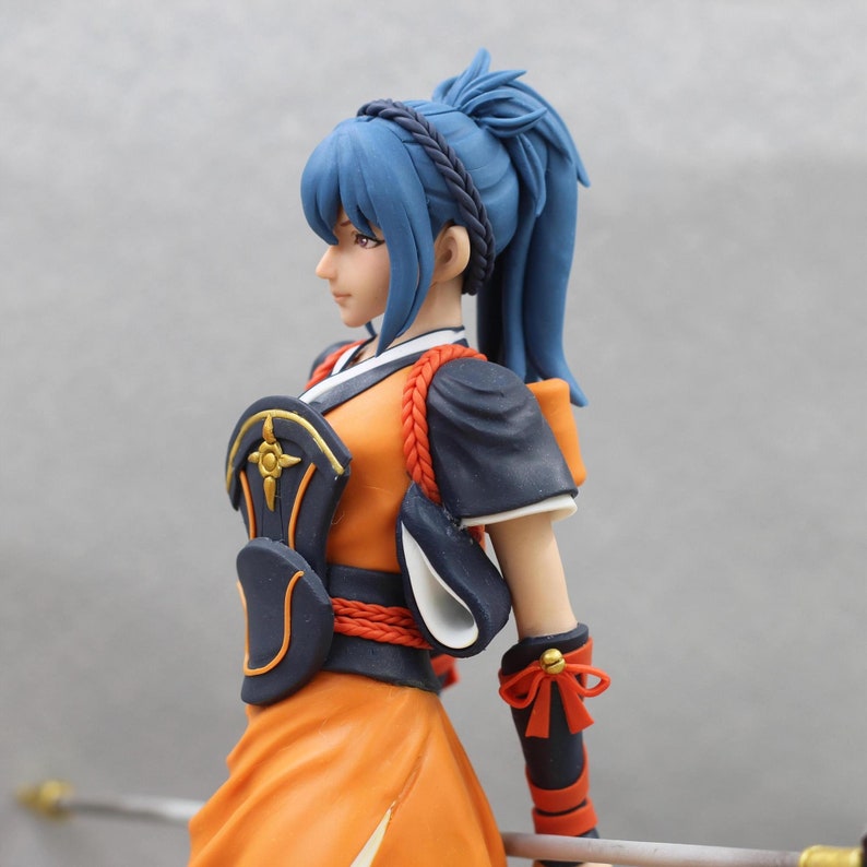 May include: A blue-haired figurine wearing a black and orange outfit with a gold and black chest piece. The figurine has a ponytail and is holding a sword.