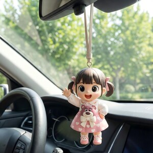 Puede incluir: Un adorno colgante para coche con una pequeña muñeca de pelo castaño en coletas, que lleva una camisa blanca de manga larga y un vestido rosa con corazones blancos y un gráfico de oso. La muñeca sonríe, saluda con una mano y cuelga de una correa.