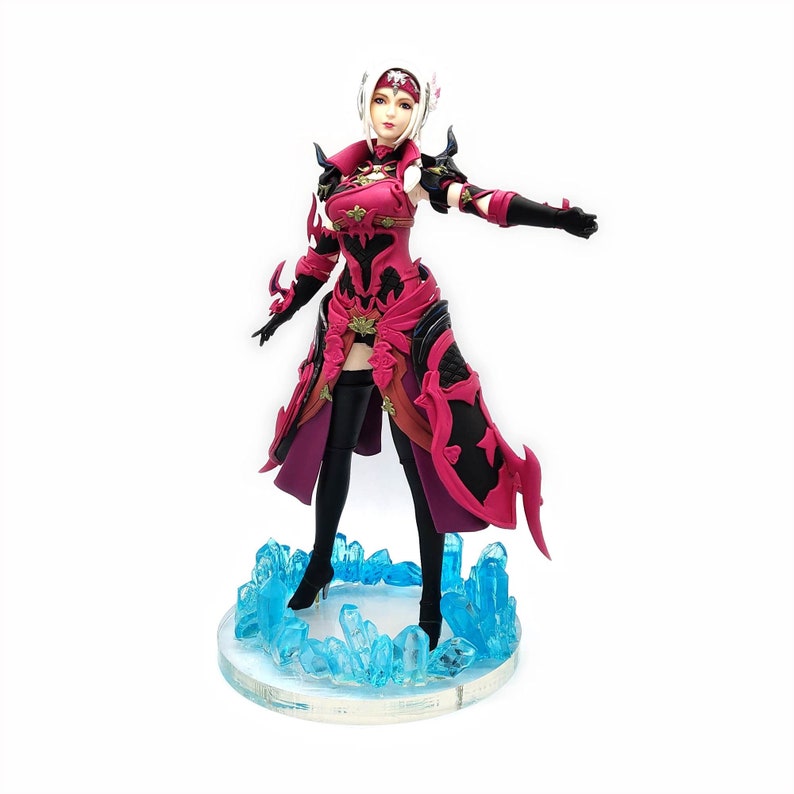 Peut inclure: Une figurine d'action rouge et noire repr&eacute;sentant un personnage f&eacute;minin aux cheveux blancs et une couronne. Elle est debout sur une base de cristal bleu.