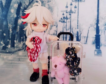 May include: A white doll with red and white hair is holding a red and white teddy bear. The doll is wearing a white shirt with a red and white pattern. The doll is standing next to a clear suitcase with a pink teddy bear and a black and white patterned fabric inside.