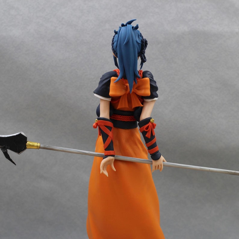 May include: A blue-haired anime figurine wearing an orange and black outfit with a large bow. The figurine is holding a long, silver spear.