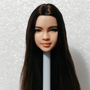 Puede incluir: Una cabeza de muñeca con cabello largo y castaño oscuro. La muñeca tiene rasgos realistas, incluidos ojos marrones, una nariz definida y una boca ligeramente entreabierta.