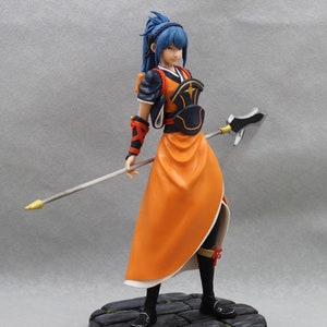 May include: A blue-haired figurine of a woman wearing a black and orange outfit with a gold and silver spear. The figurine is standing on a stone base.