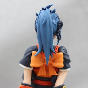 May include: A blue haired doll with a large orange bow tied around her neck. The doll is wearing a dark blue and red outfit.