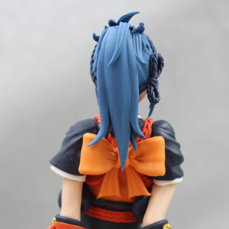 May include: A blue haired doll with a large orange bow tied around her neck. The doll is wearing a dark blue and red outfit.