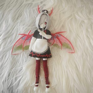 May include: A doll with white hair and red and black striped stockings wearing a black and white dress with a white apron and red wings. The doll is wearing a nurse's hat with a red cross.