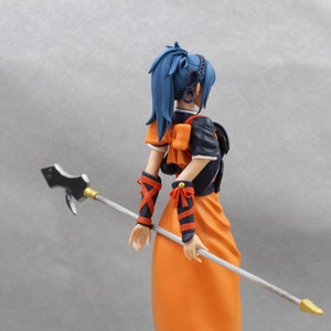 May include: A blue-haired anime figurine holding a silver and gold spear. The figurine is wearing a black and orange outfit with a large orange bow.