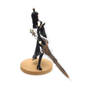 Peut inclure: Une figurine noire et blanche d'une femme en robe longue noire tenant une grande &eacute;p&eacute;e &agrave; lame blanche et brune. La figurine est pos&eacute;e sur une base ronde en bois.