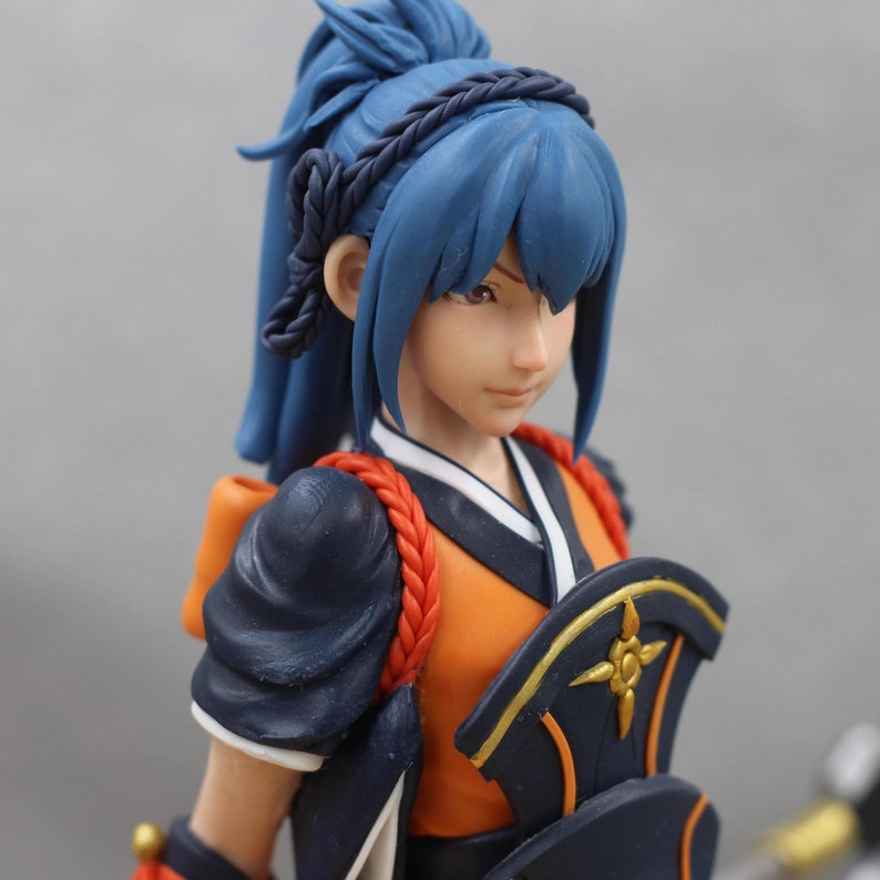 May include: A blue-haired figurine with a black braid, wearing a dark blue and orange outfit with gold accents. The figurine has a serious expression and is looking to the right.