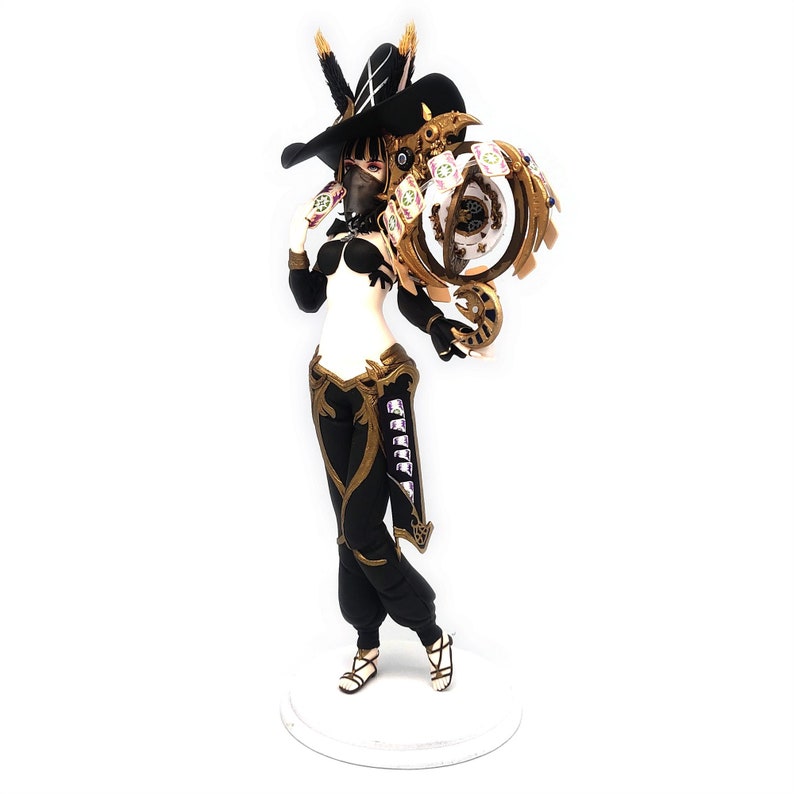 Peut inclure: Une figurine noire et dor&eacute;e repr&eacute;sentant un personnage f&eacute;minin portant un chapeau noir et un masque. Le personnage tient un objet orn&eacute; de couleur or et blanc dans sa main droite. La figurine est pos&eacute;e sur une base blanche.