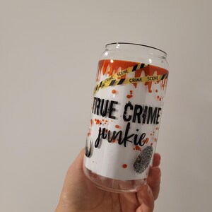 May include: A white tumbler with a black and orange design. The tumbler has the words "True Crime Junkie" and a yellow and black crime scene tape design.