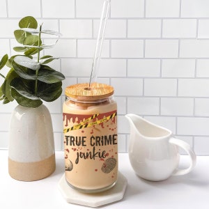 May include: A glass with a wooden lid and a straw, filled with a light brown liquid. The glass has a graphic of red blood dripping down the side with the text "TRUE CRIME junkie" and yellow crime scene tape.