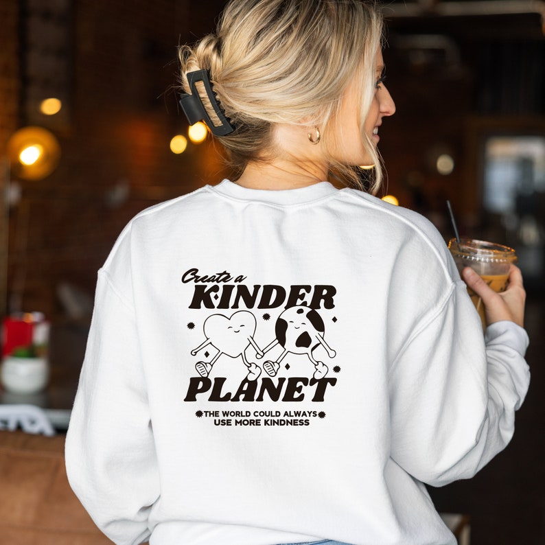 Może przedstawiać: Biała bluza z czarnym nadrukiem graficznym, na kt&oacute;rym widnieje napis "Create a Kinder Planet" z sercem i planetą trzymającymi się za ręce. Tekst pod grafiką brzmi "The world could always use more kindness."