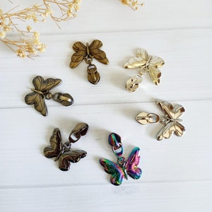 May include: A selection of butterfly-shaped zipper pulls in bronze, silver, and iridescent rainbow finishes. The zipper pulls are displayed on a white surface, with dried floral accents.