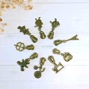 May include: A selection of antique bronze zipper pulls, each with a different charm. Charms include a teddy bear, Eiffel Tower, and a four-leaf clover. The zipper pulls are arranged on a light-colored surface. The image is taken from above.
